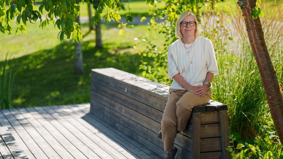 Kvinna sitter på en bänk utomhus med grönska runt om sig och tittar in i kameran.