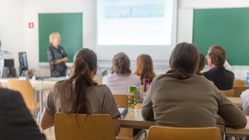 Föreläsningssal på universitet med studenter i förgrunden och en lärare vid tavlan.