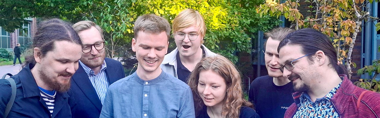 En grupp personer står utomhus framför en byggnad och tittar tillsammans på en laptop. De ser glada och engagerade ut, och omgivningen är grön med träd och buskar i bakgrunden.