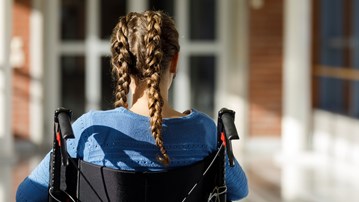 Flicka med flätor sitter i rullstol framför byggnad.