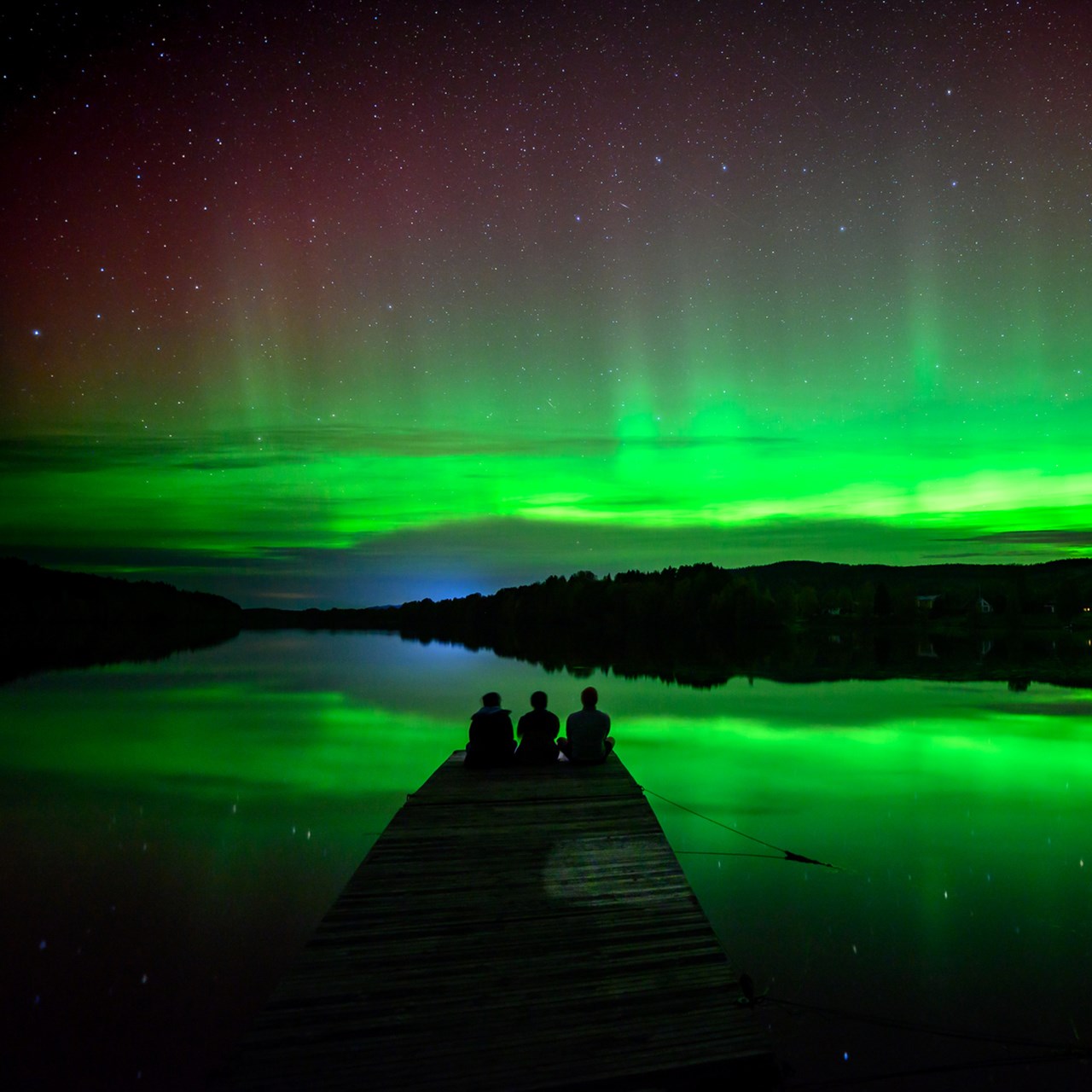 A group of people sitting on a dock watching the aurora reflecting in a river.
