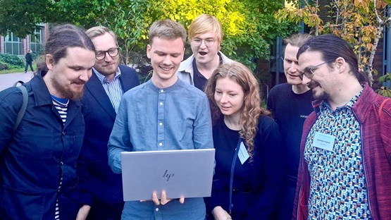 En grupp personer står utomhus framför en byggnad och tittar tillsammans på en laptop. De ser glada och engagerade ut, och omgivningen är grön med träd och buskar i bakgrunden.