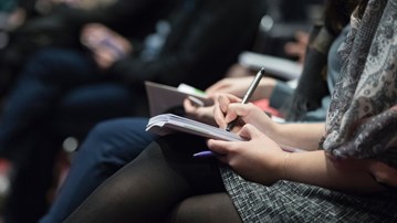 Närbild på en kvinna som sitter med block och penna på seminarium och antecknar.