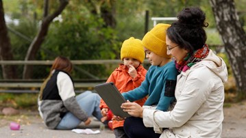 Förskolepedagog tittar på en surfplatta med barn utomhus på en förskola.