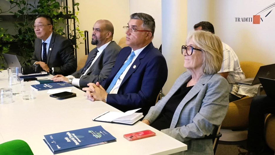 Four people in siuts sit by a table and listen to a presentation. At the table are research folders, notebooks and glasses with water.