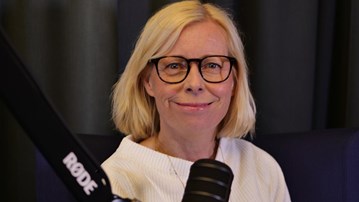 Kvinna med blont hår och glasögon sitter i en fåtölj, med en podmic i förgrunden.