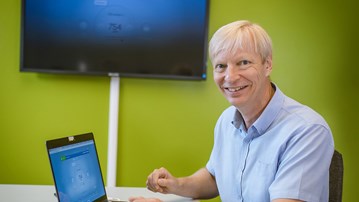 Kary Främling, professor vid institutionen för datavetenskap, sitter vid skrivbord med en dator och en skärm på väggen